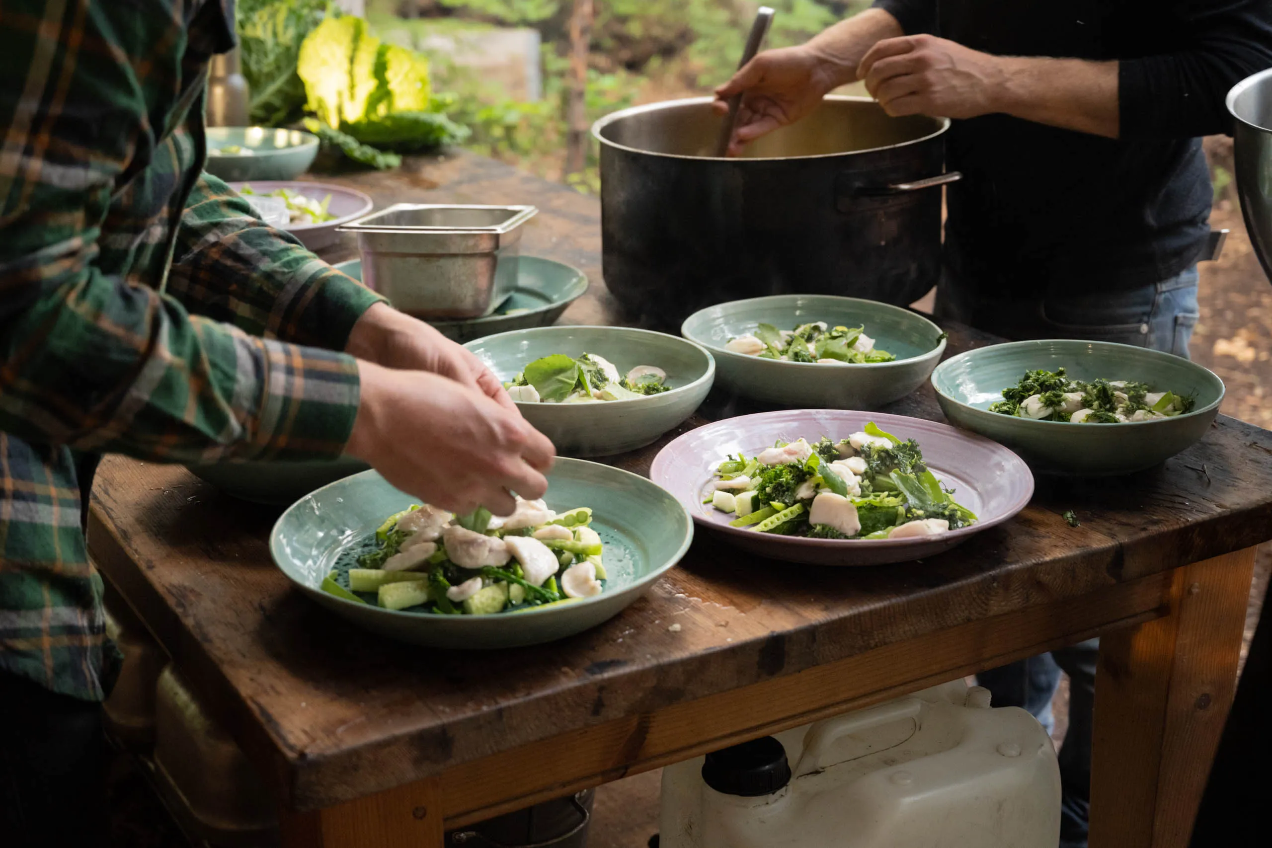 Two cooks plate green vegetable dishes into ceramic bowls at a rustic wooden prep table, one stirring a large pot in the background.