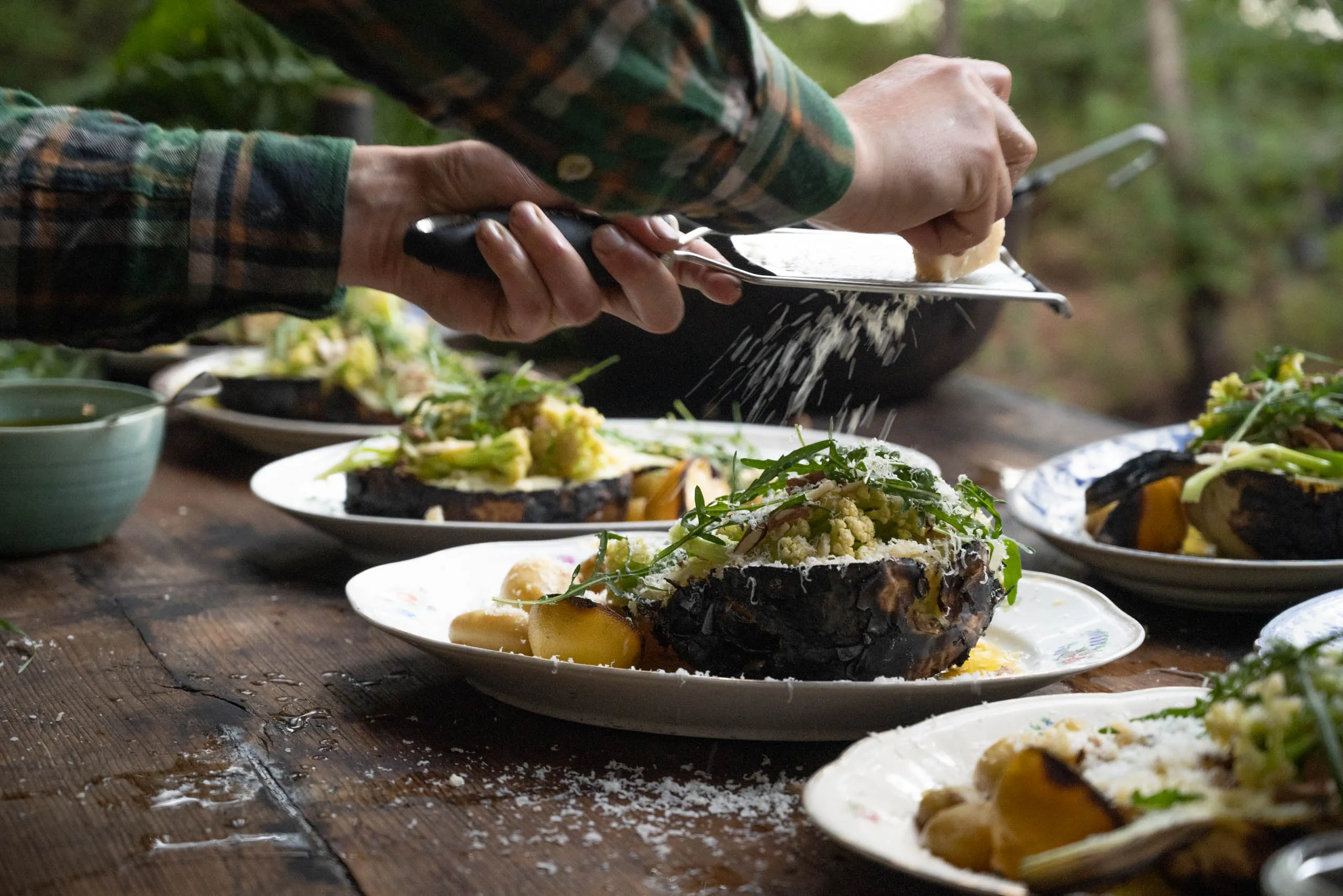 Hands grate fresh cheese over plates of charred grilled cauliflower with rocket and shaved vegetables on a forest dinner table.