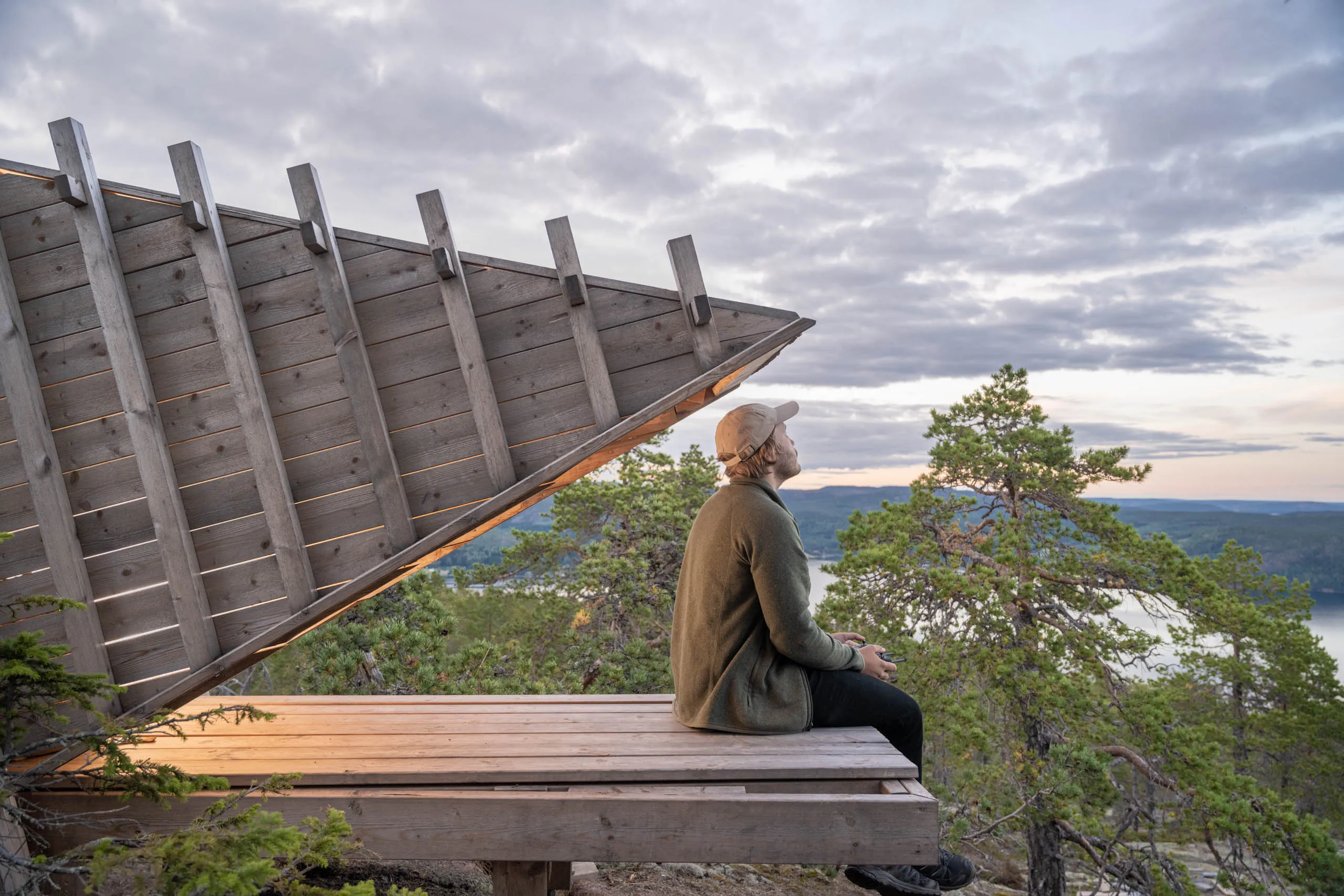 A person in an olive sweater and cap sits on the wooden bench of a slanted timber lookout shelter on the High Coast of Sweden, looking out over a fjord at dusk.