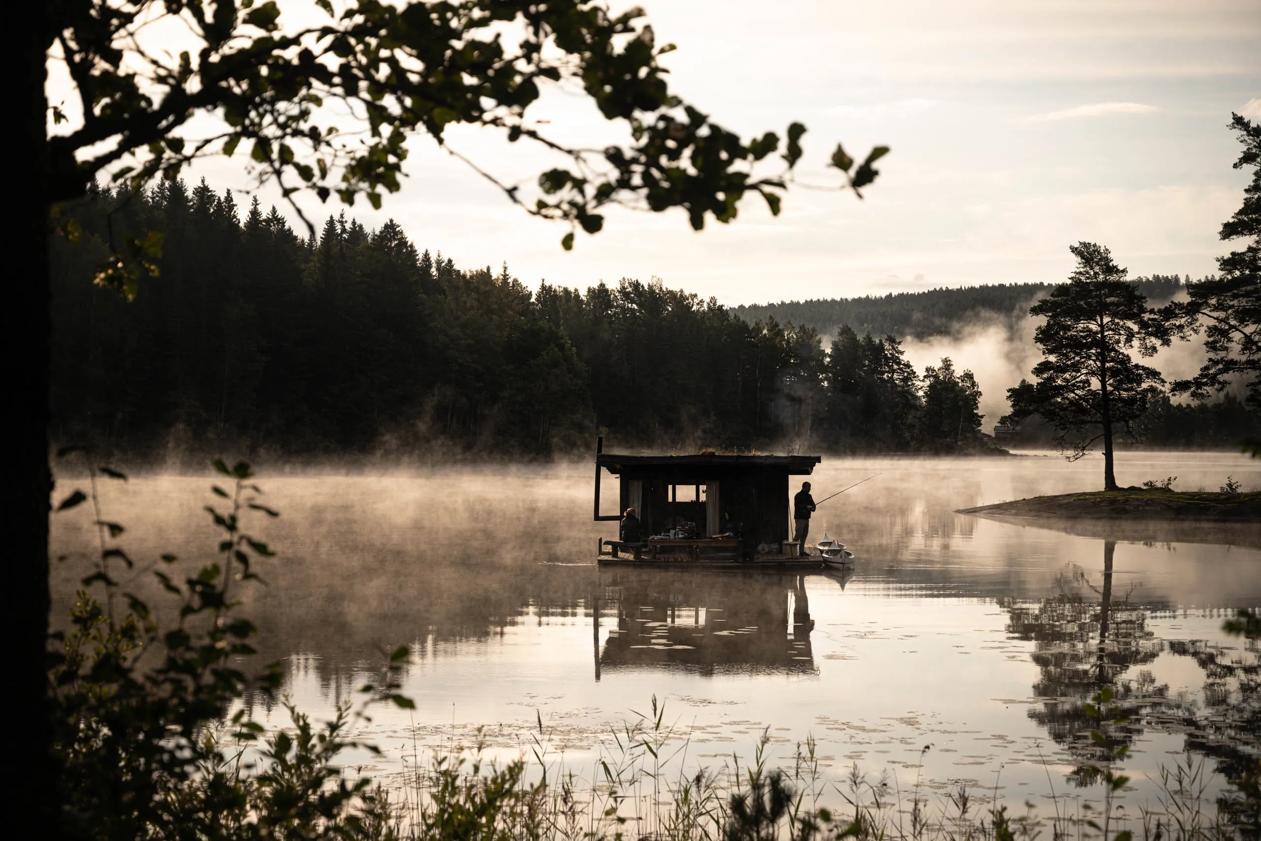 Silhouette of a person fishing from the deck of a floating sauna-cabin on a Swedish lake at dawn, mist rising off the still water.