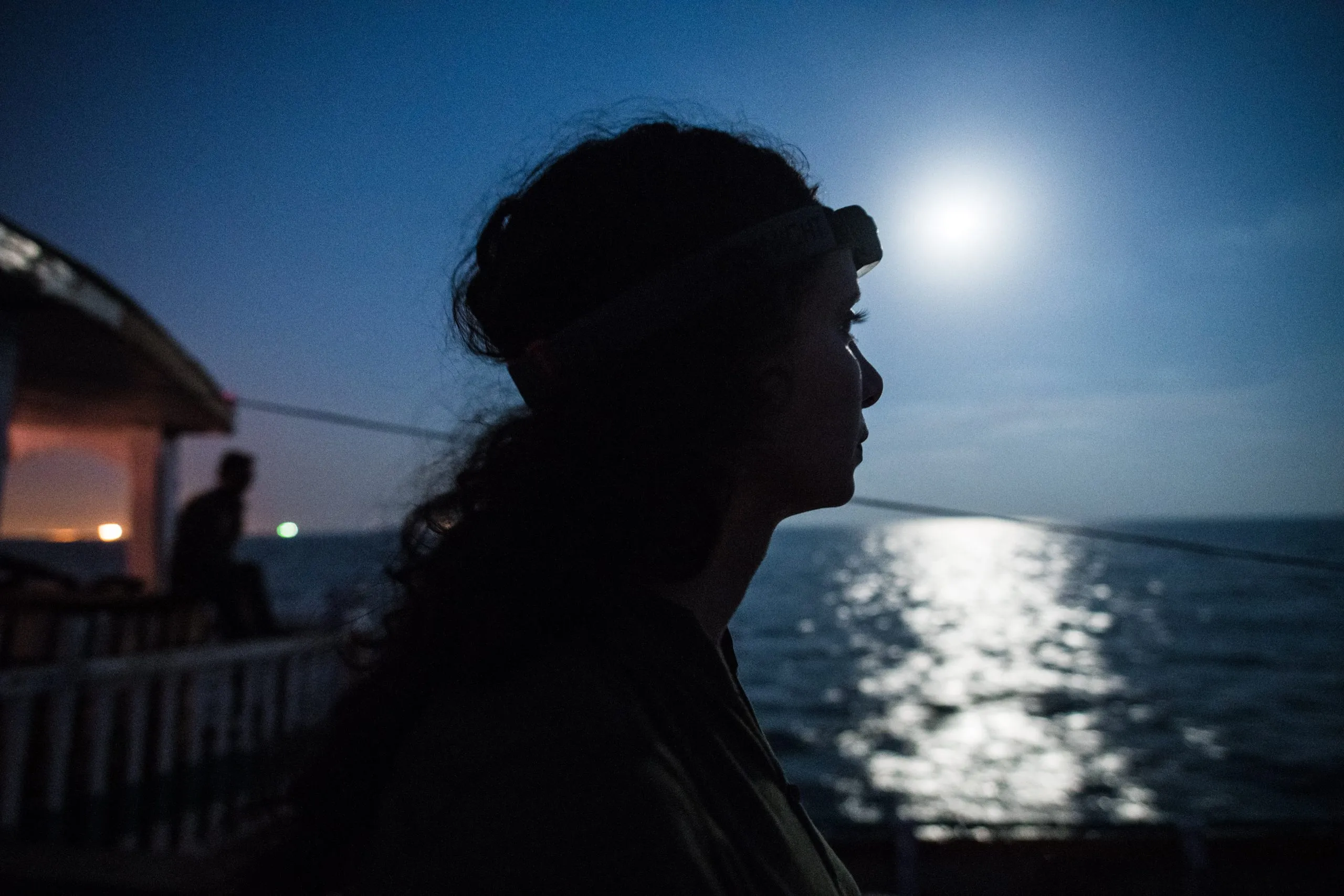 Silhouette of a traveler with a headlamp on her forehead, looking out across the Arabian Sea as the moon rises over the horizon.