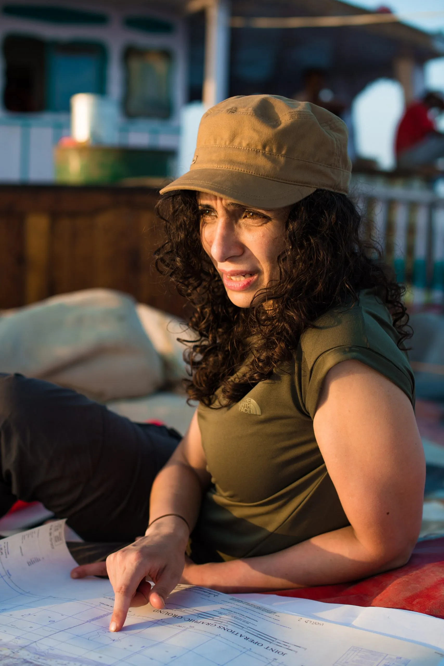 A traveler in an olive shirt and tan cap studies a Joint Operations Graphic marine chart laid out on the dhow's deck in golden afternoon light.