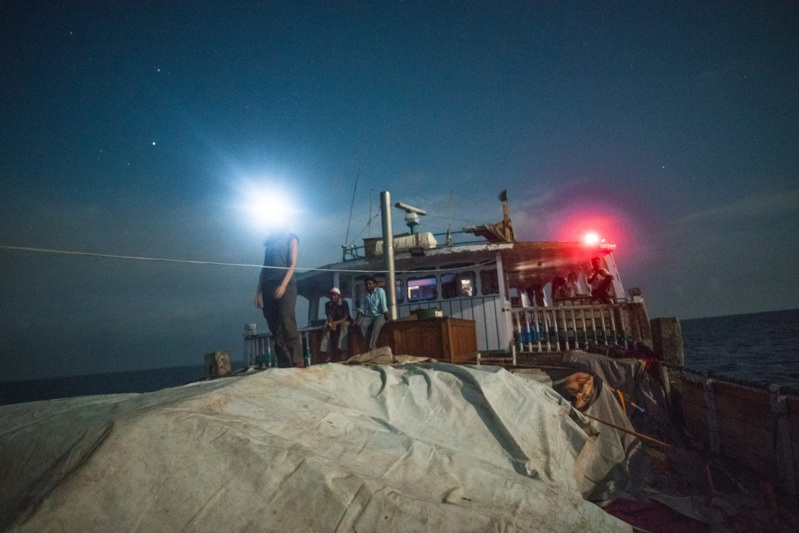 Crew and passengers on the deck of the cargo dhow at night under the stars, a person standing with a bright headlamp, red navigation light glowing on the cabin.