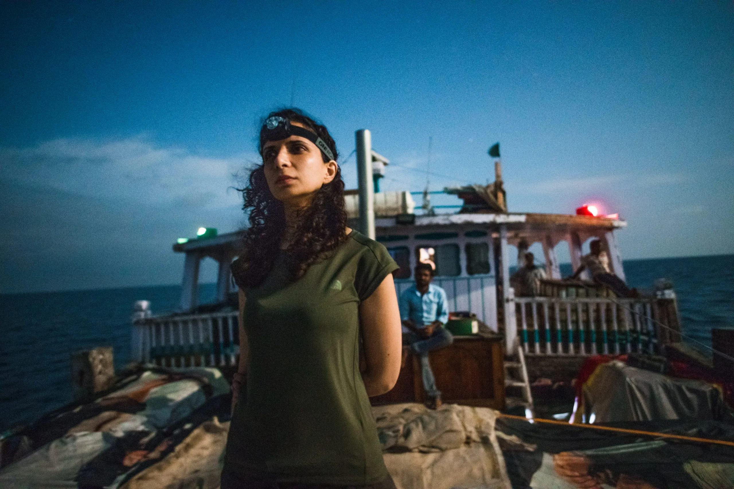 A traveler with a headlamp on her forehead stands on the dhow's deck at twilight, the wooden wheelhouse and crew visible behind her.