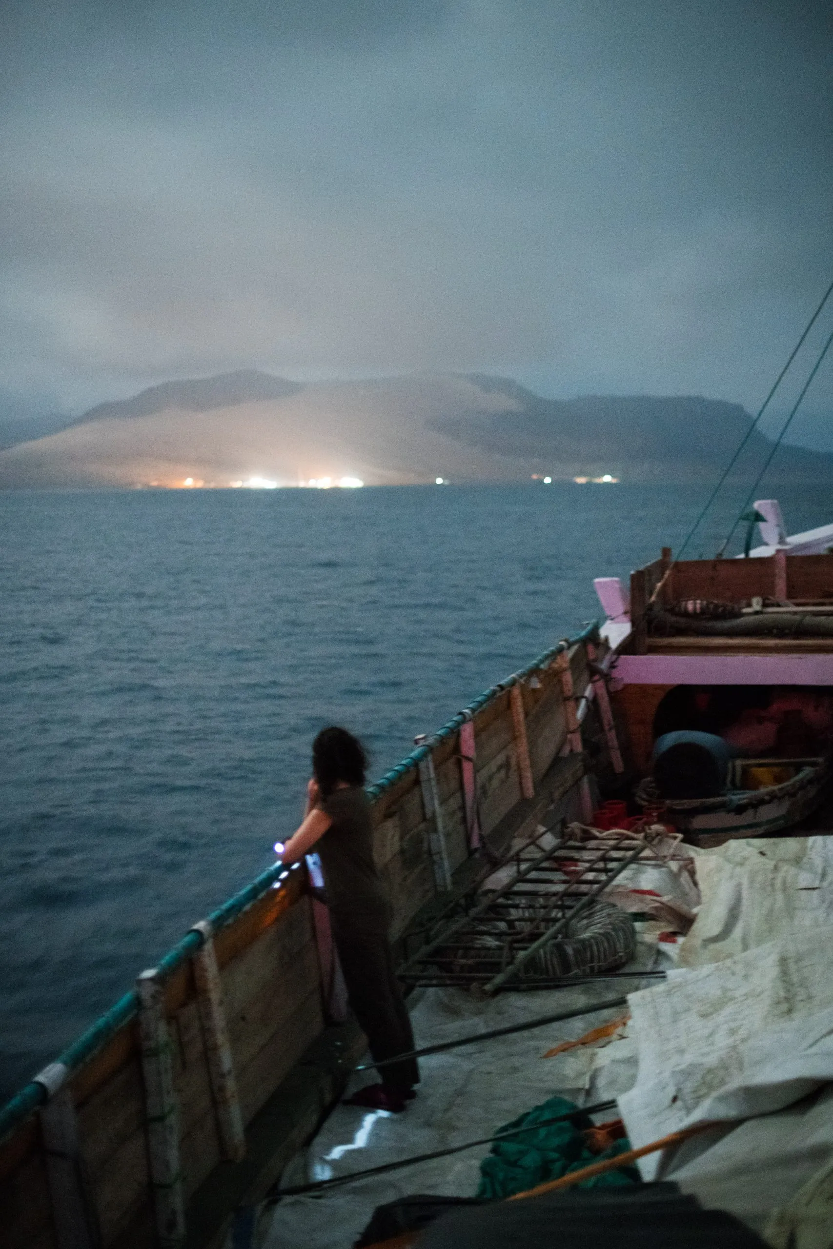 A traveler at the rail of the dhow looks toward the lights of Hadibu glowing under the misty silhouette of Socotra's mountains as the boat approaches at night.