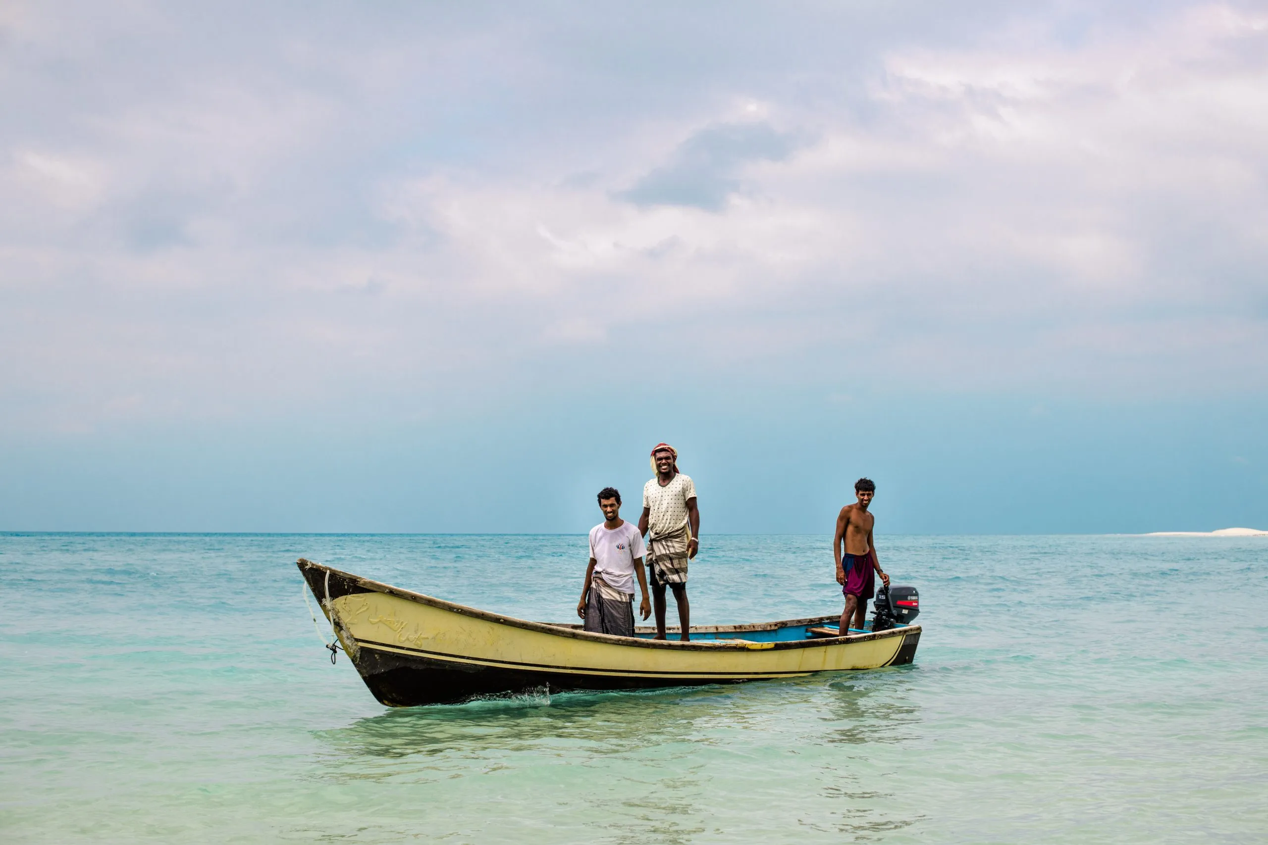 Three Socotri fishermen stand on a small yellow wooden boat in clear shallow turquoise water off Socotra's western coast.
