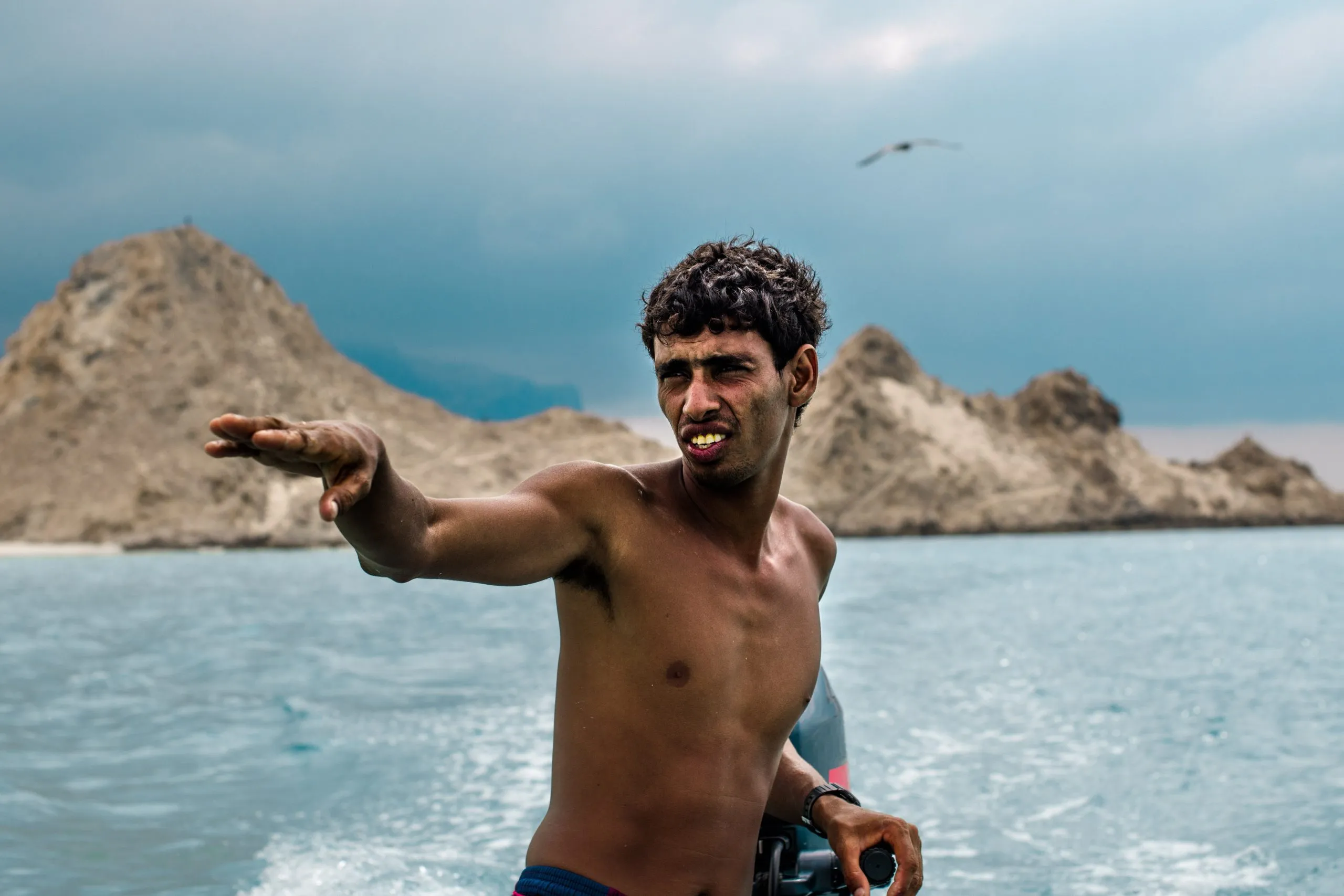 A young Socotri fisherman gestures with an outstretched arm from a small boat, jagged sand-and-rock peaks rising behind him on the coast.