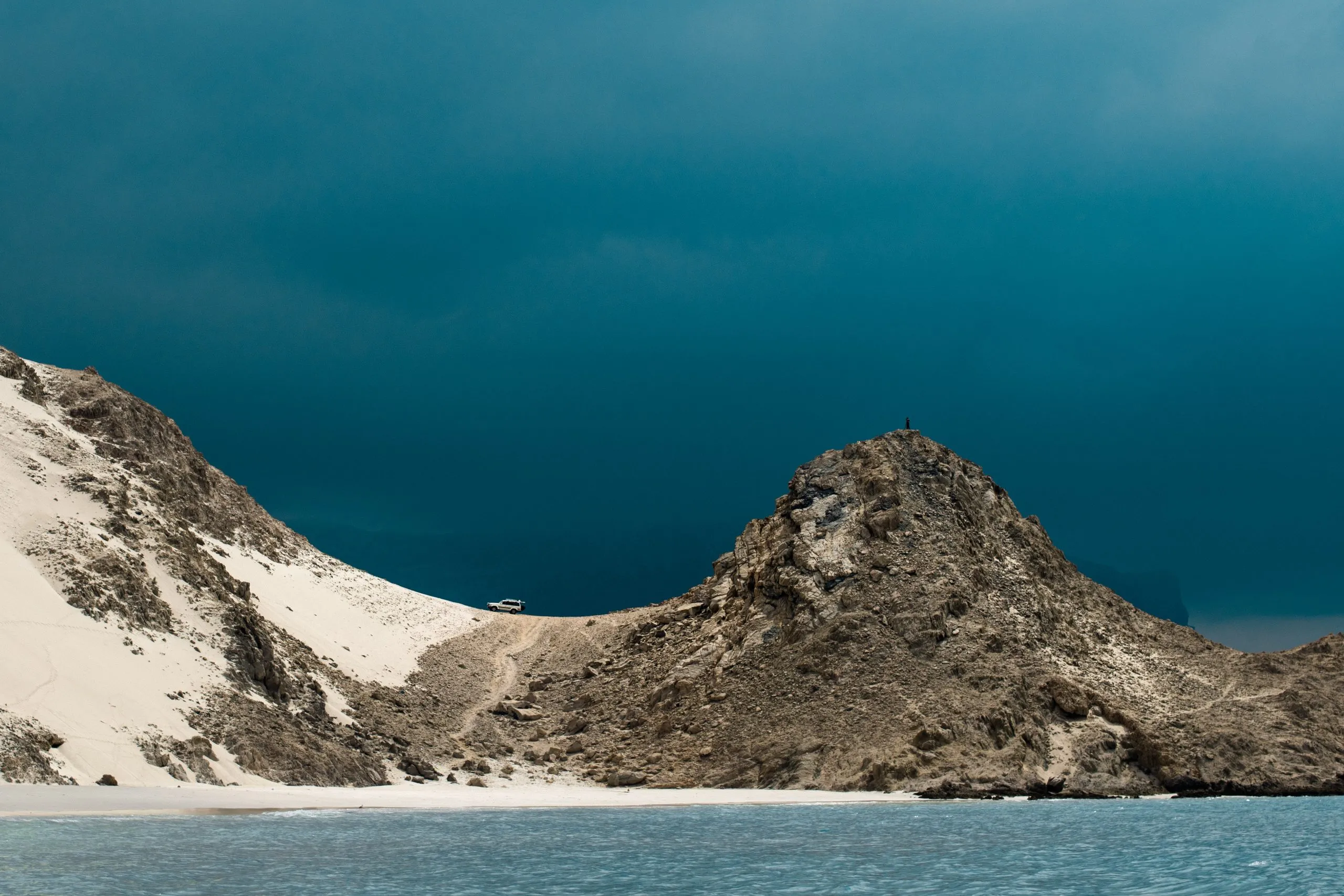 A pale dune sweeps up between dark rocky peaks against a deep teal sky, a tiny figure visible on top of the rock.