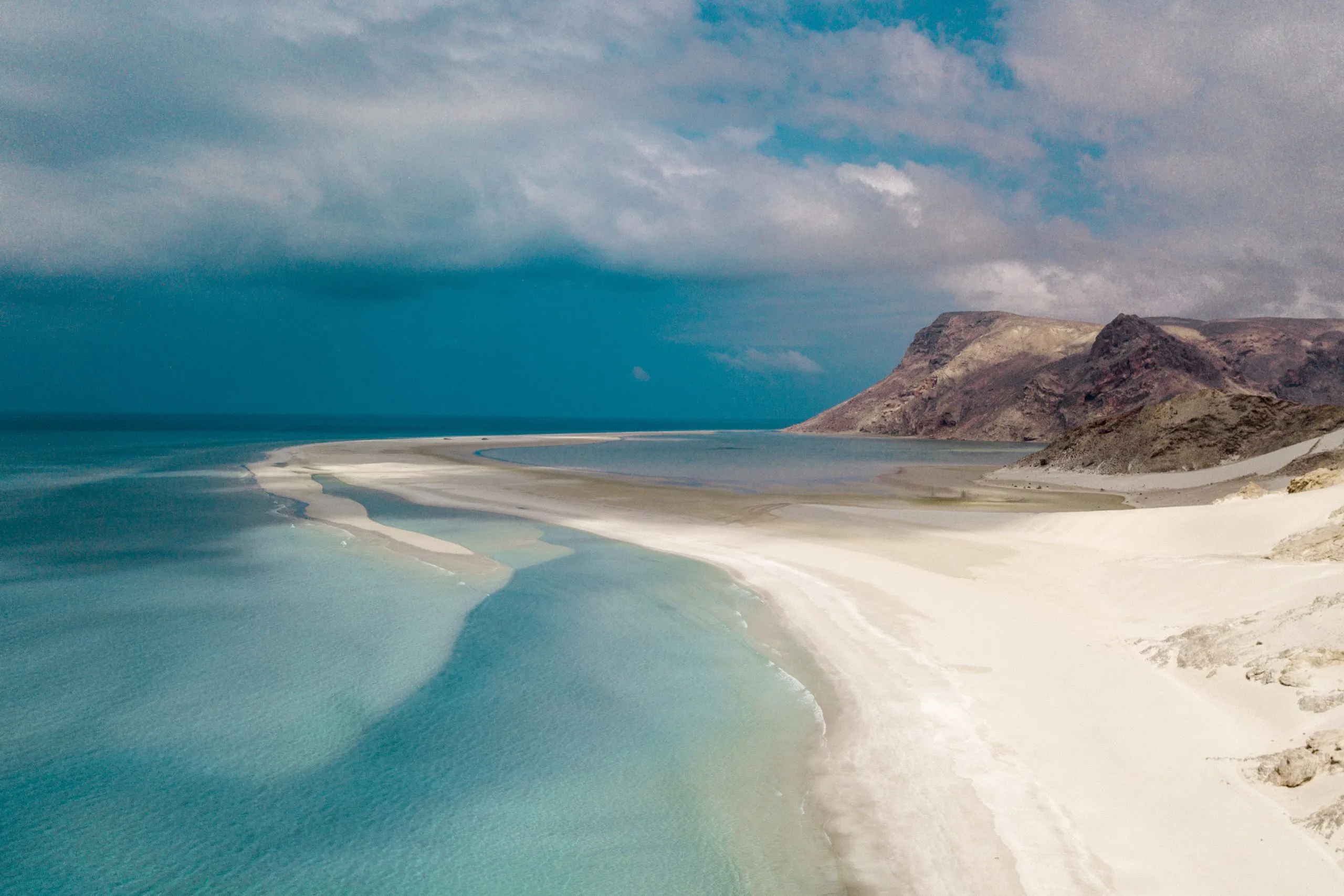 Aerial view of a long white sandbar curving between turquoise lagoon water and rocky desert hills at Detwah Lagoon, Socotra.