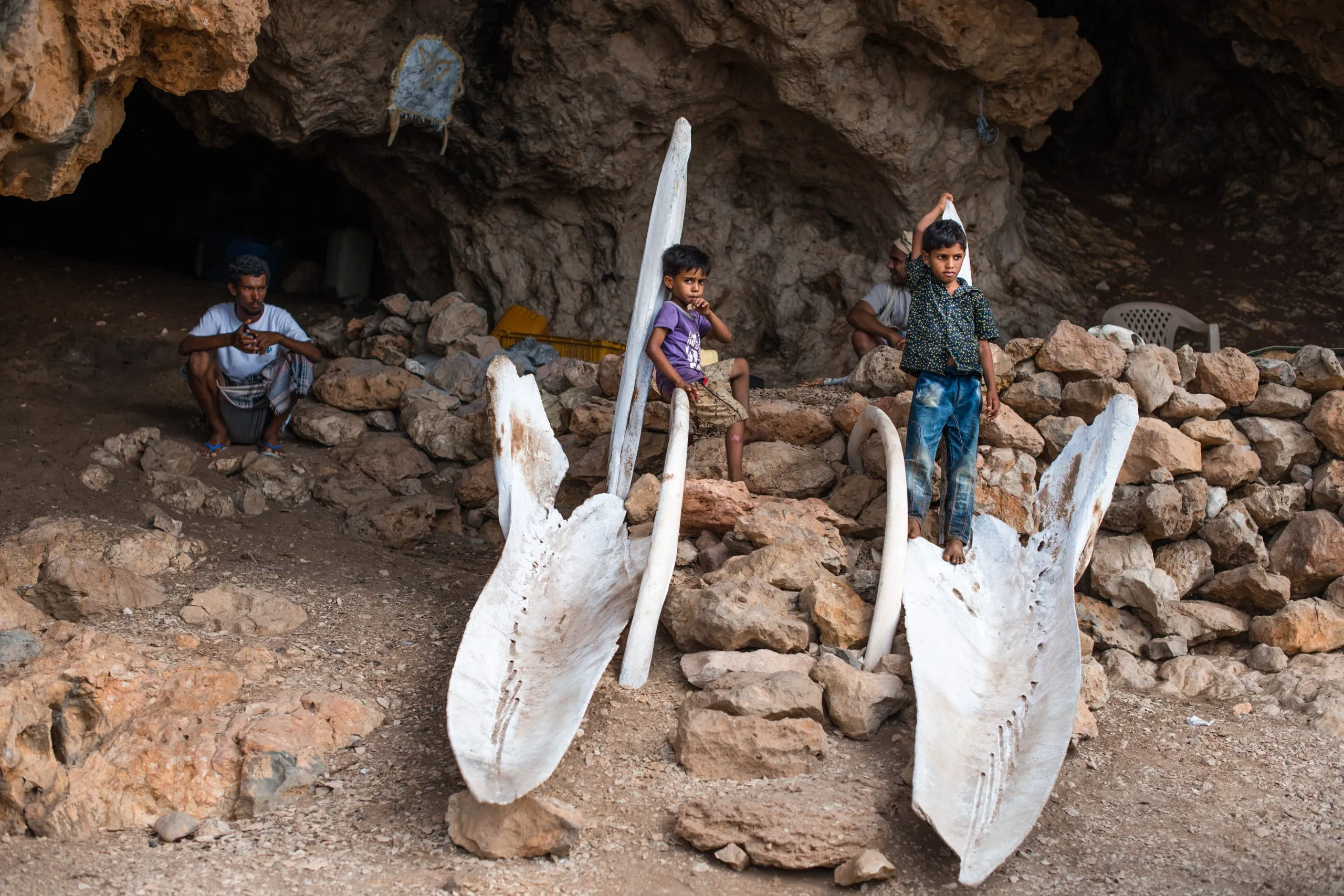 Two Socotri children stand on enormous whale jaw bones inside a rocky cave shelter, an older man seated to the side.