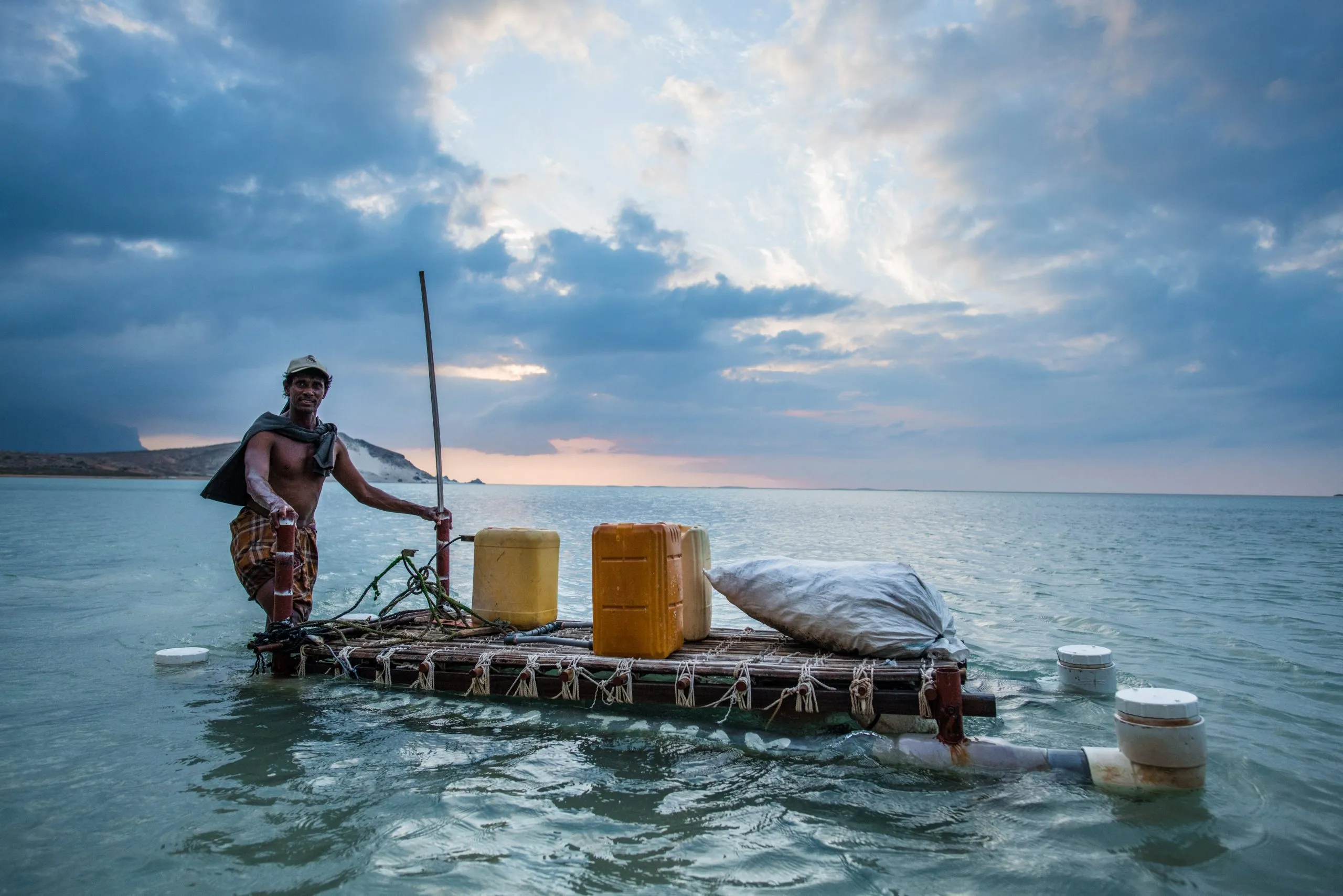A Socotri man stands waist-deep beside a small wooden raft loaded with yellow jerry cans at sunset, pole in hand.