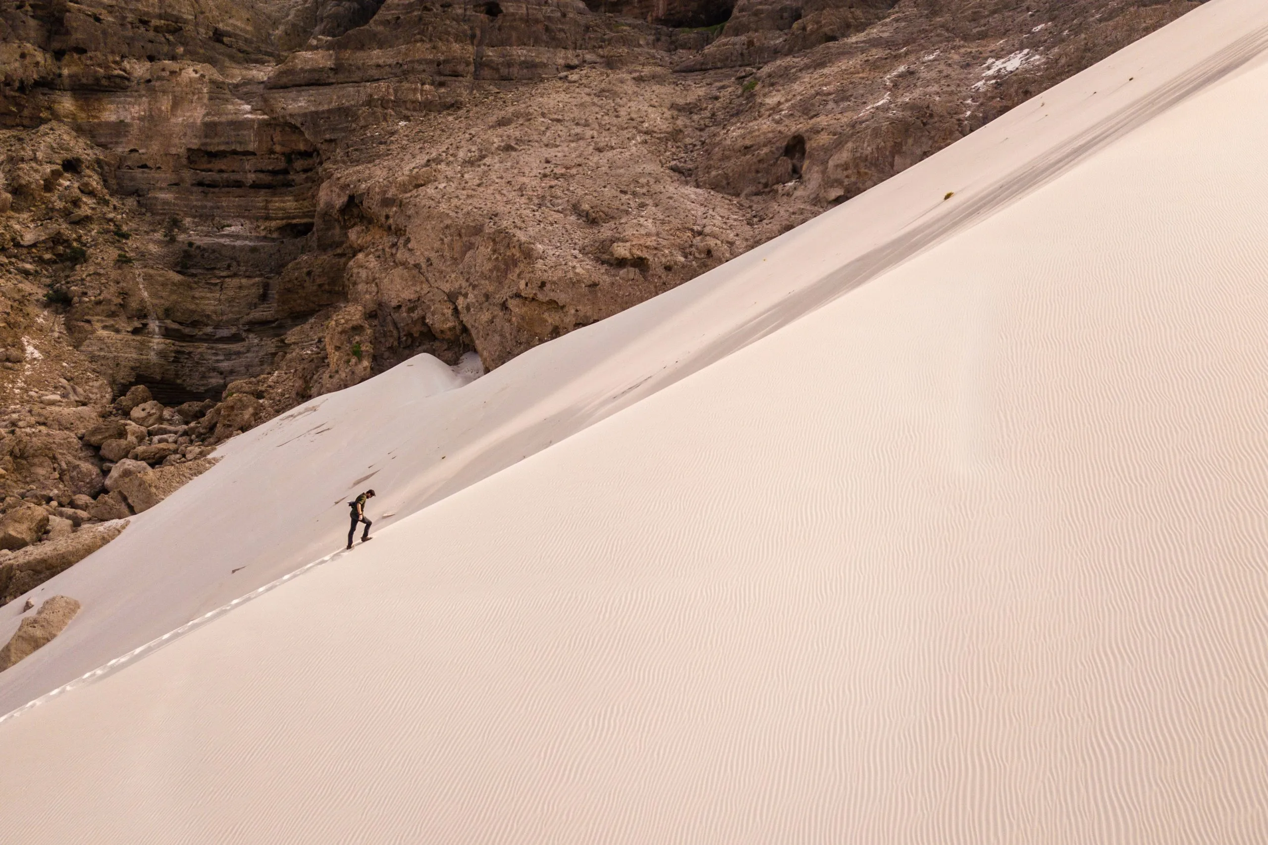 A tiny figure climbs a steep pale dune wedged at the foot of a banded canyon wall on Socotra.