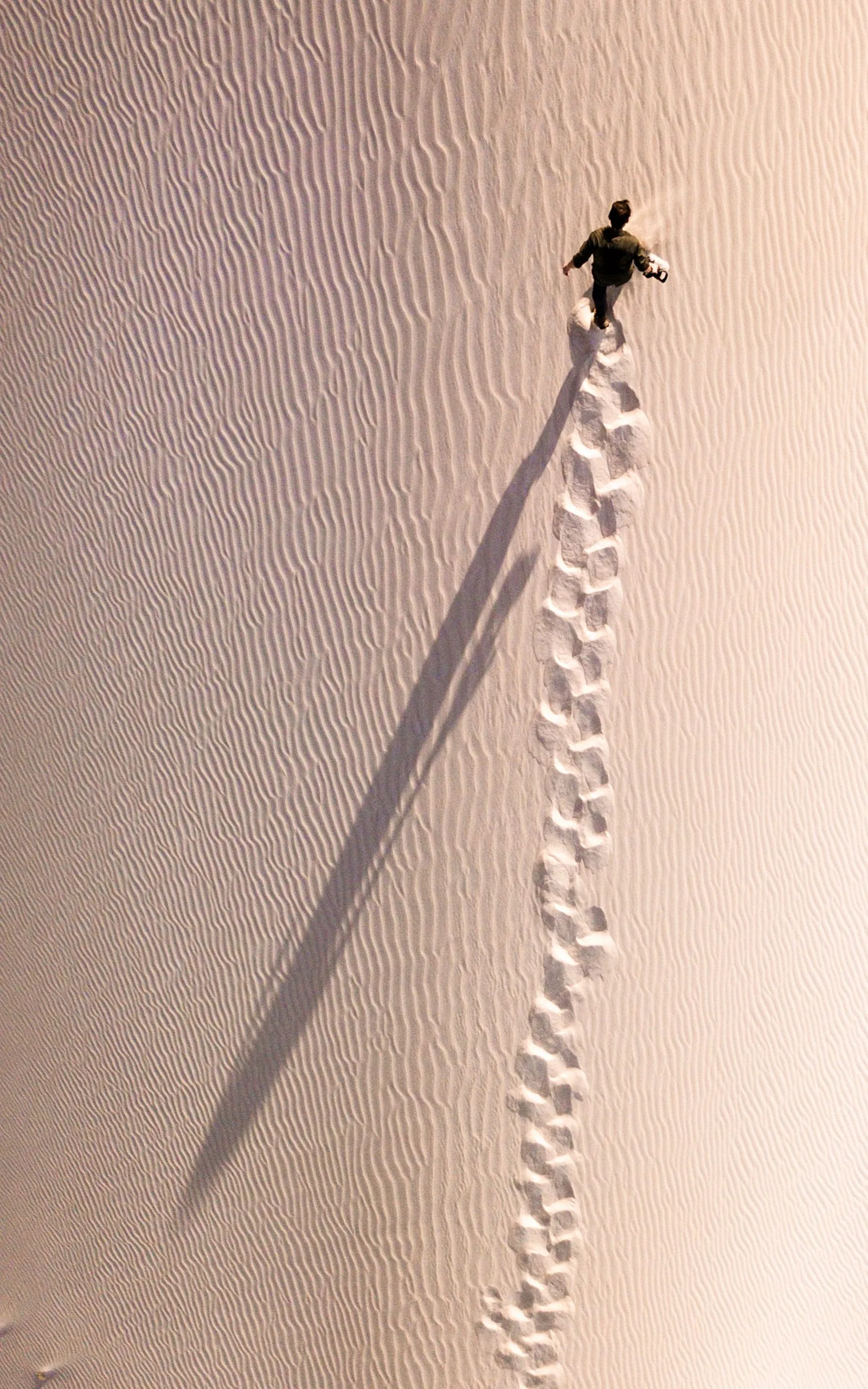 Aerial view of a person walking up a sand dune in golden side-light, a long shadow and a deep trail of footprints behind them.