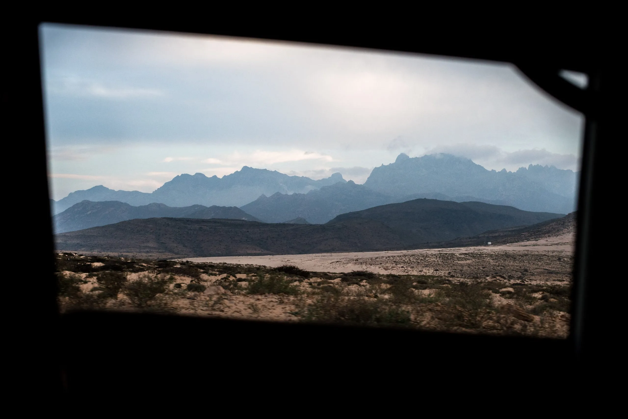 Through a vehicle window, the jagged silhouette of the Haghier Mountains rises in layers of blue at dusk on Socotra.