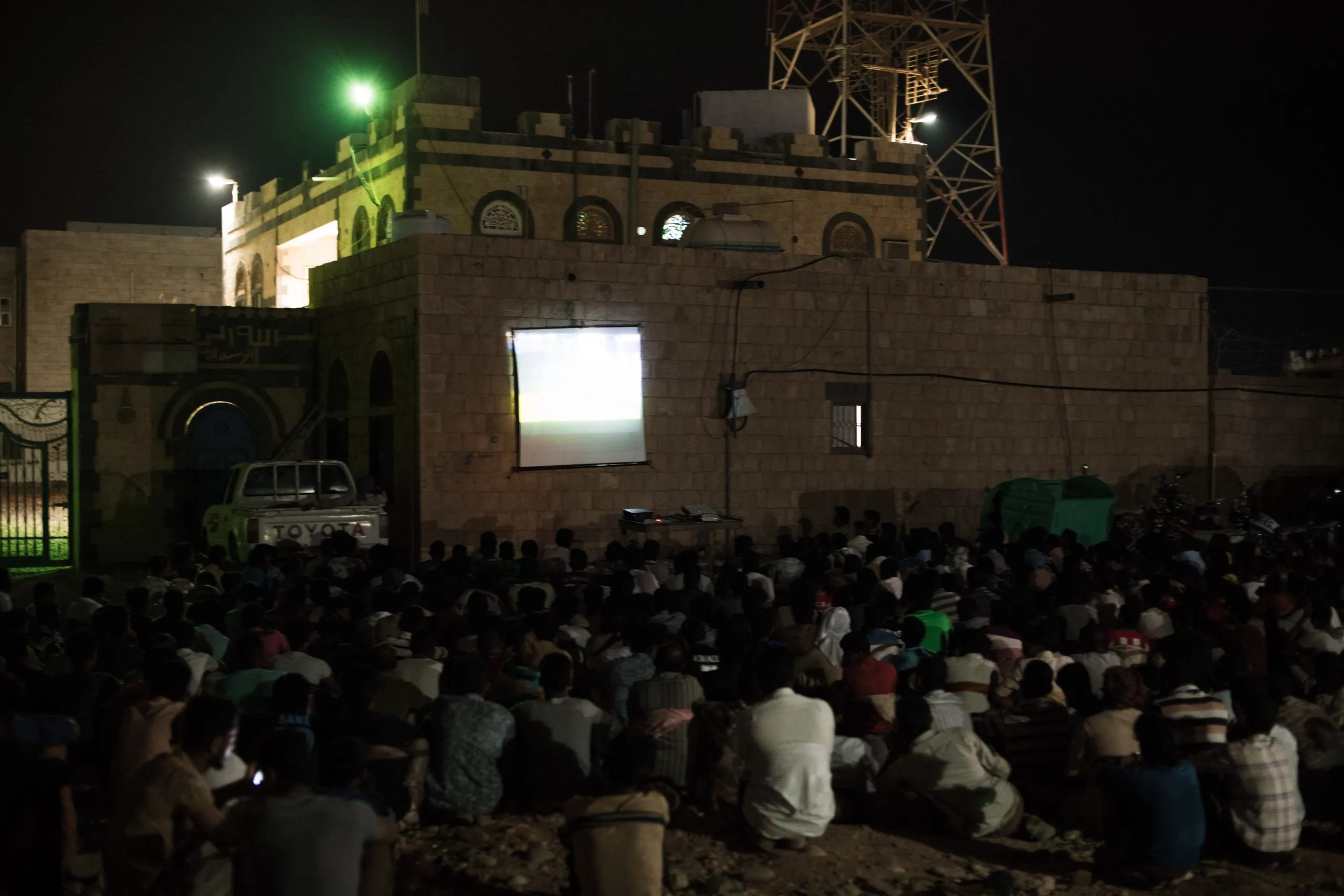 A large crowd in Hadibu watches an outdoor film projected onto the wall of a stone building at night, a Toyota truck parked nearby.