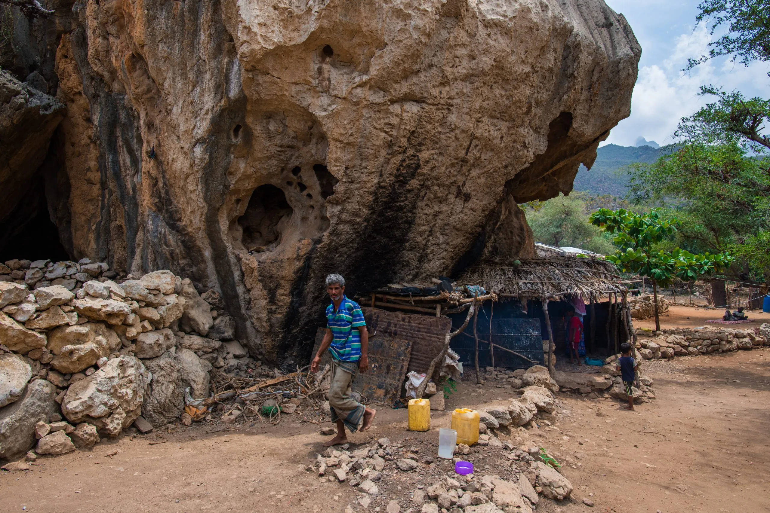 A Socotri man steps out of a rock-shelter dwelling tucked under a deeply weathered limestone overhang, jerry cans on the dirt floor and a palm-thatched lean-to built against the rock.