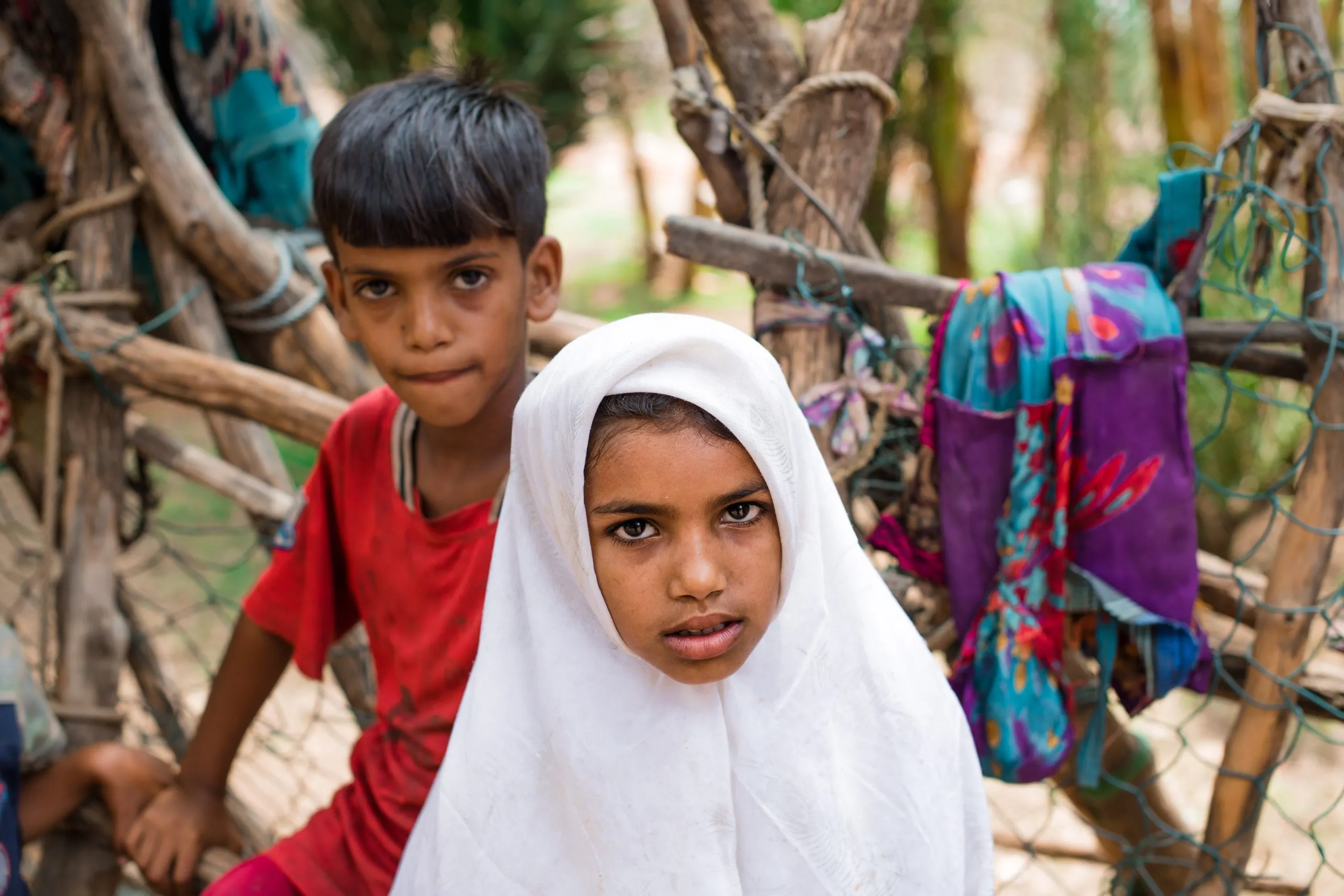 Two Socotri children — a boy in a red shirt and a girl in a white headscarf — look at the camera in front of a wooden shelter draped with colourful fabrics.