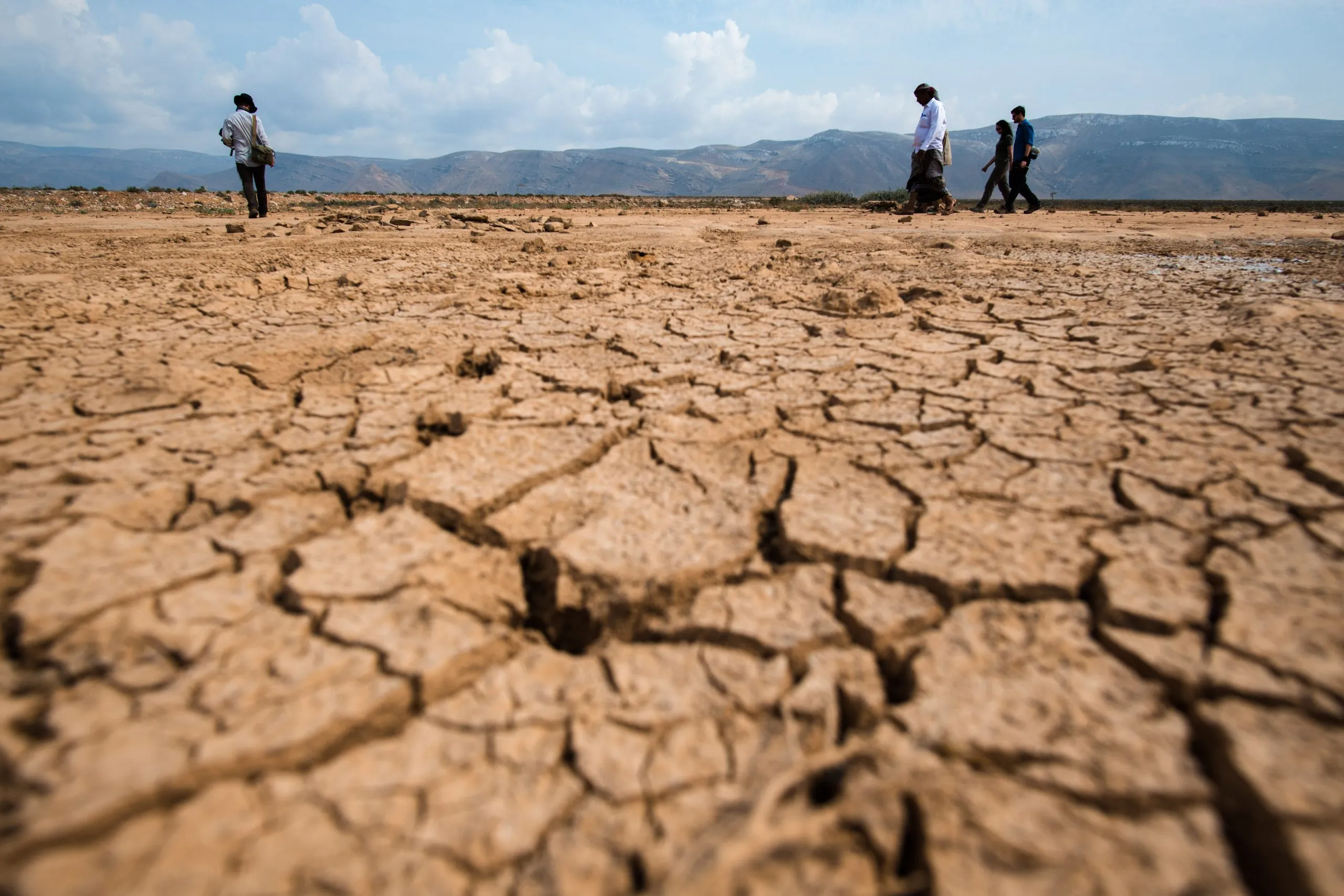 Cracked dry mud fills the foreground of a parched lake bed on Socotra, a small group walking across it in the distance with mountains behind.