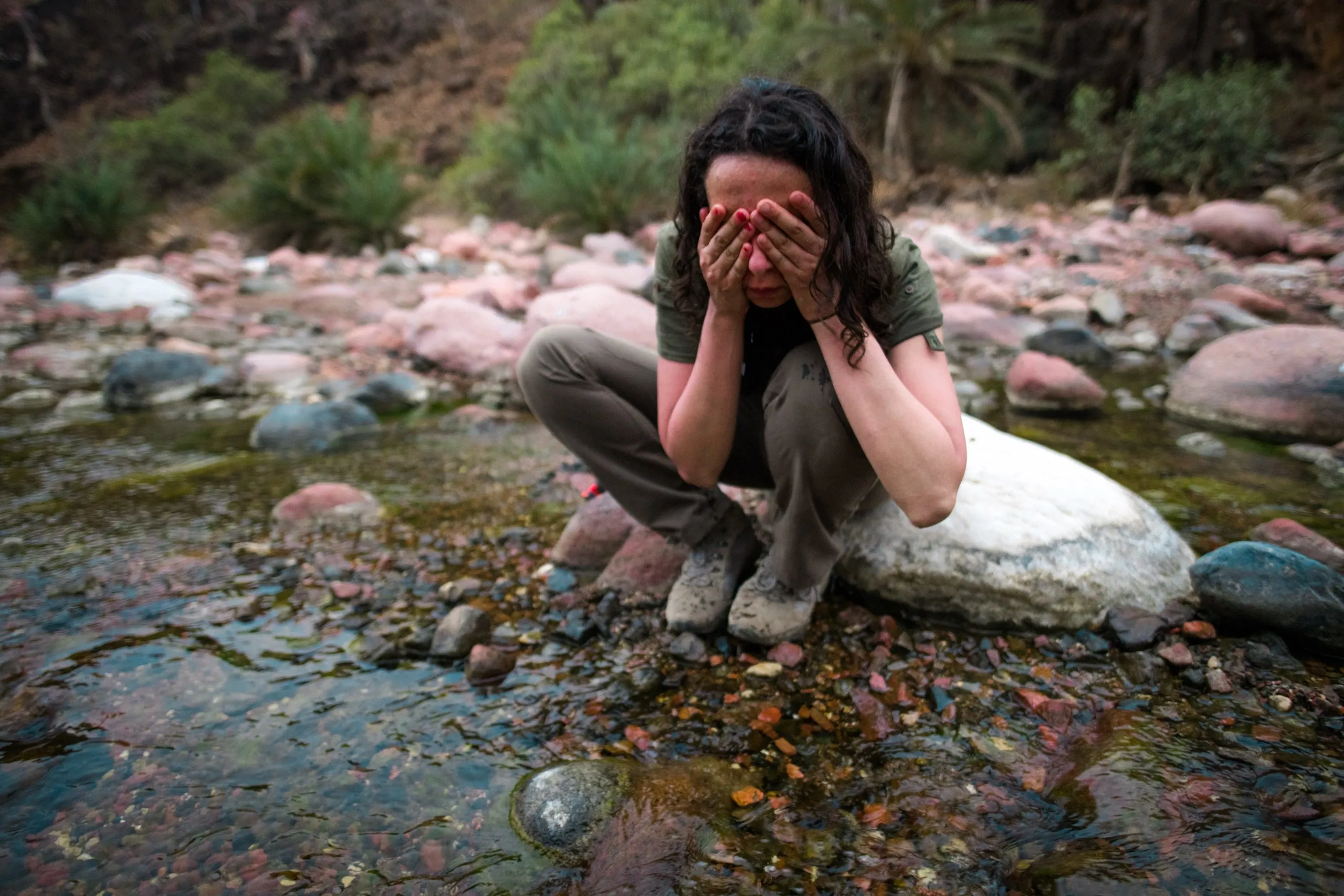 A traveler crouches by a clear stream in a wadi to wash her face, surrounded by colourful rounded river stones and palms.