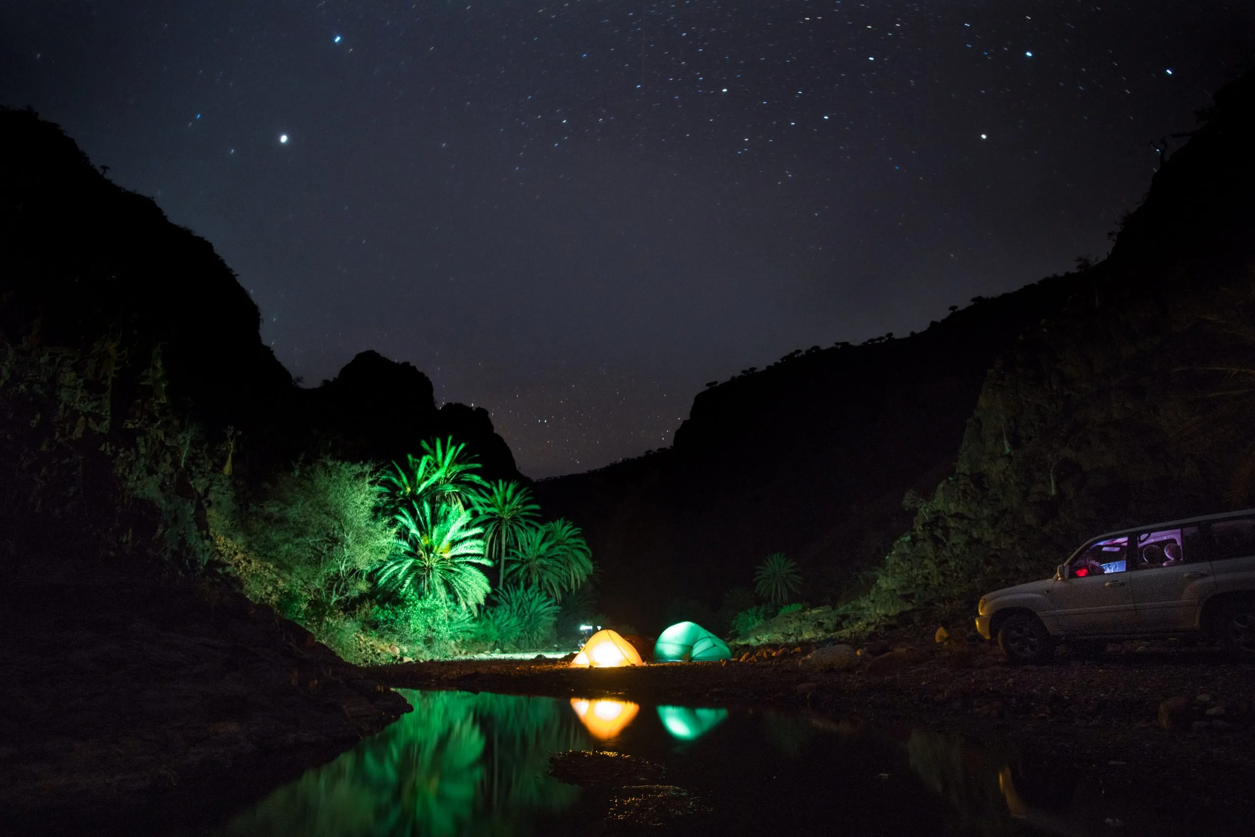 A wadi camp by night under a starry sky on Socotra: two glowing tents pitched beside a still pool, palms uplit in green and a parked Land Cruiser to one side.