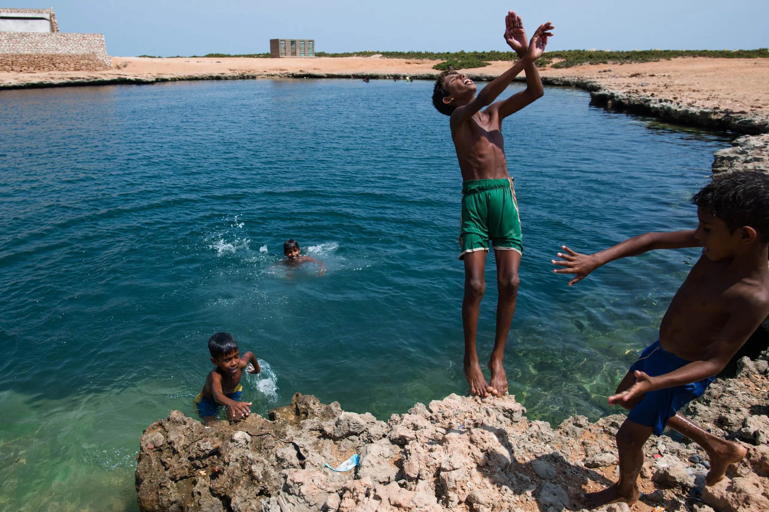 Socotri boys leap and swim in a clear blue freshwater spring on the coast, one boy caught mid-air above the rocks.